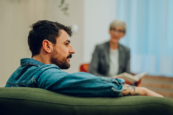 Man raising newborn alone in therapy session, looking thoughtful while woman in background holds a notebook and listens. Man raising newborn alone in therapy session, looking thoughtful while woman in background holds a notebook and listens.