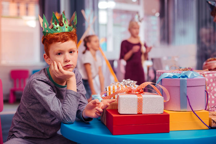 Sad boy wearing a crown sitting by birthday presents, reflecting exclusion and family conflict at a child's party. Sad boy wearing a crown sitting by birthday presents, reflecting exclusion and family conflict at a child's party.