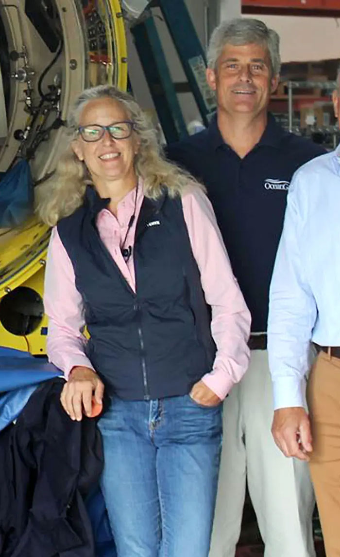 OceanGate CEO’s wife smiling beside OceanGate team members in a workshop setting with submersible equipment nearby. OceanGate CEO’s wife smiling beside OceanGate team members in a workshop setting with submersible equipment nearby.