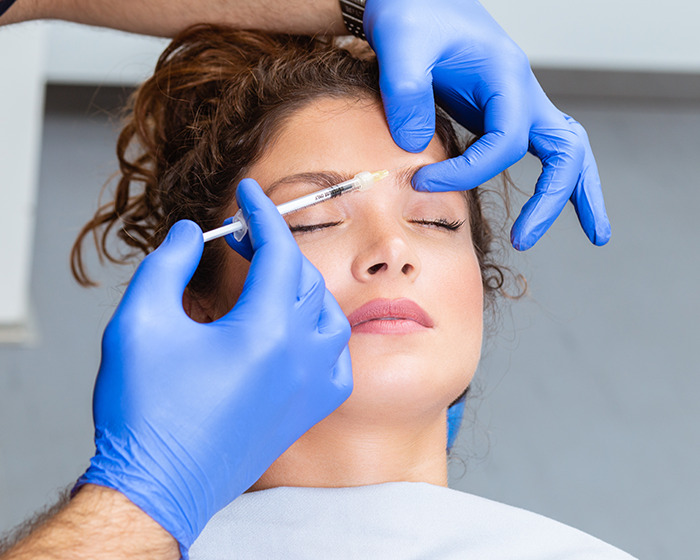 Woman receiving cosmetic injection to forehead by plastic surgeon wearing blue gloves in clinical setting Woman receiving cosmetic injection to forehead by plastic surgeon wearing blue gloves in clinical setting