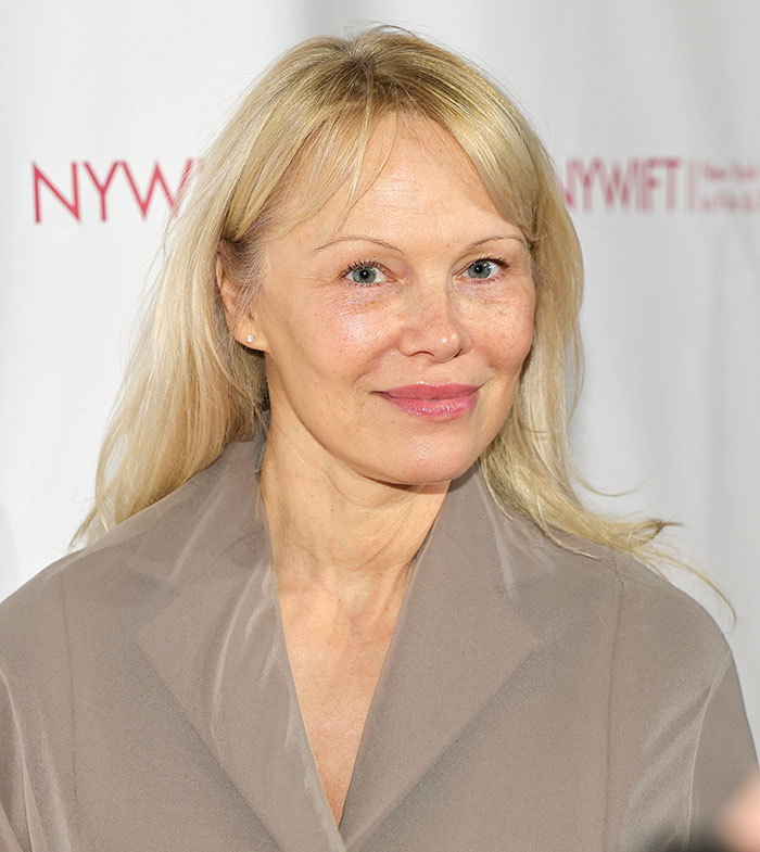 Pamela Anderson smiling with natural look and minimal makeup at an indoor event with blonde hair and beige coat. Pamela Anderson smiling with natural look and minimal makeup at an indoor event with blonde hair and beige coat.