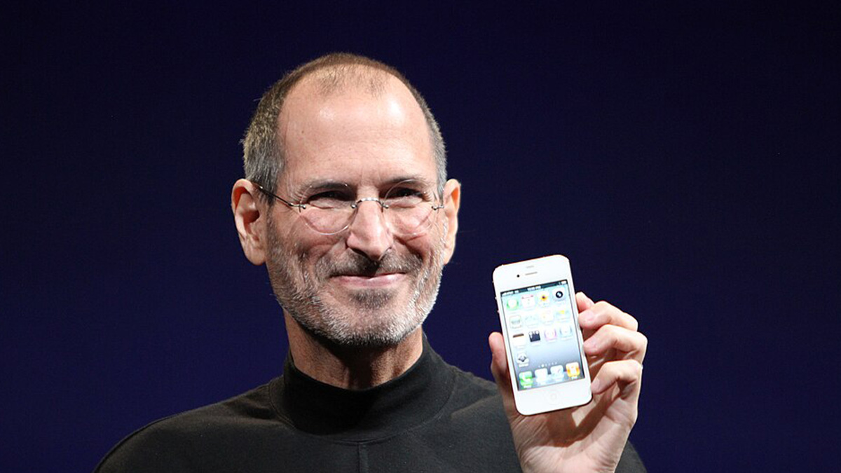 Steve Jobs holding an iPhone, smiling during a product launch event against a dark background.