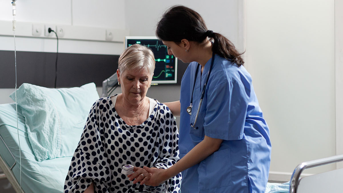 Nurse comforting elderly patient in hospital room with medical equipment monitoring vital signs nearby.