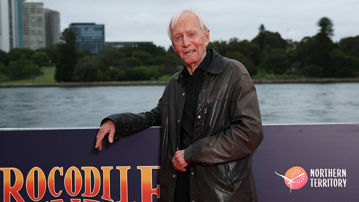 Paul Hogan posing outdoors by water near a Crocodile Dundee sign, wearing a black leather jacket and shirt.