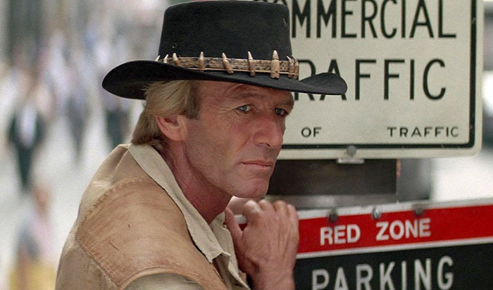 Paul Hogan as Crocodile Dundee wearing a black hat with teeth details, standing near a commercial traffic sign. Paul Hogan as Crocodile Dundee wearing a black hat with teeth details, standing near a commercial traffic sign.