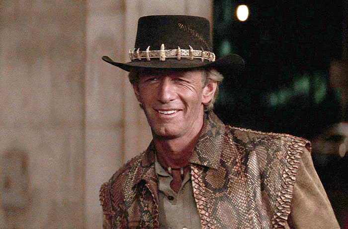 Paul Hogan as Crocodile Dundee wearing a crocodile tooth hat and snakeskin vest, smiling in a movie scene. Paul Hogan as Crocodile Dundee wearing a crocodile tooth hat and snakeskin vest, smiling in a movie scene.