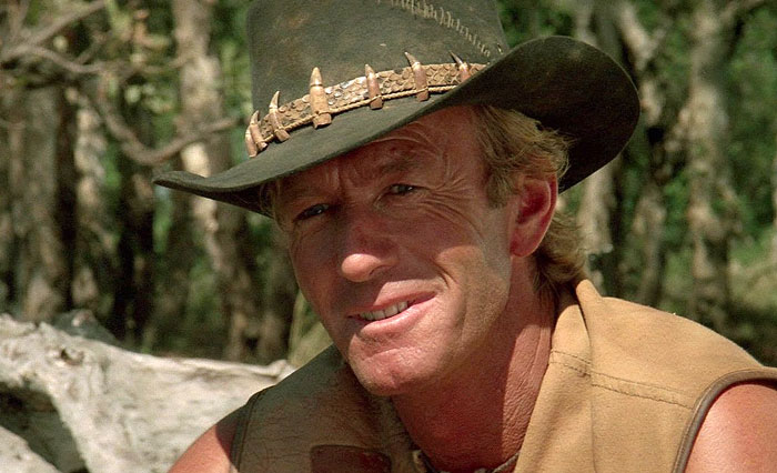 Paul Hogan as Crocodile Dundee wearing a hat with teeth, smiling in an outdoor forest setting. Paul Hogan as Crocodile Dundee wearing a hat with teeth, smiling in an outdoor forest setting.