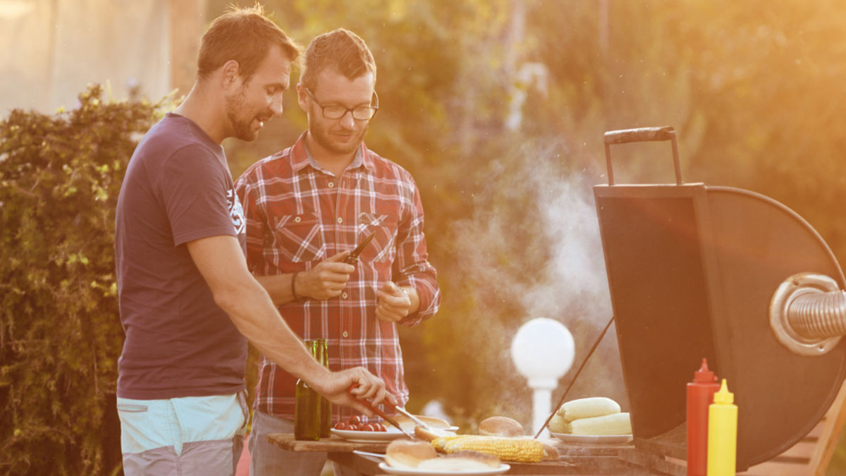 Two men grilling corn and hot dogs outside on a sunny day with a smoky barbecue grill and condiments nearby