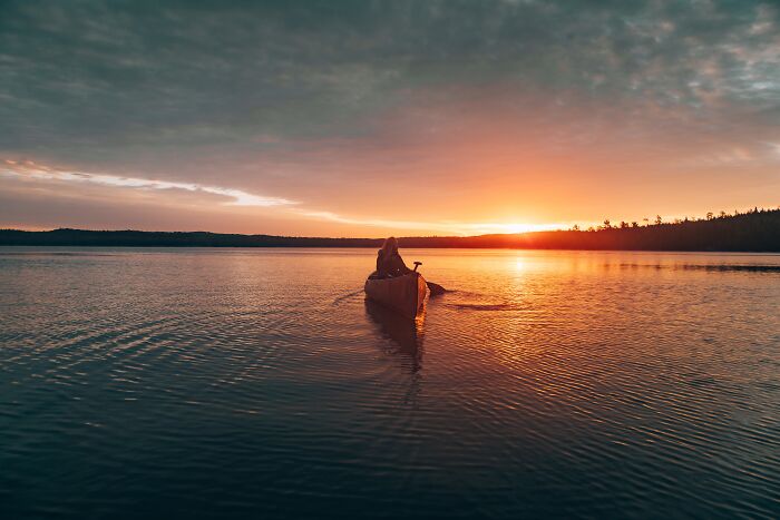 Person in a canoe paddling on a calm lake at sunset, evoking a spooky and mysterious vibe of time travel and resurrection.