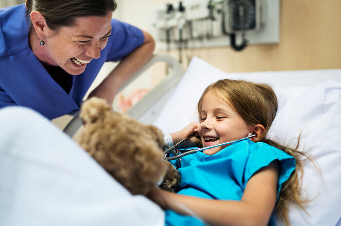 Nurse smiling at a young girl in a hospital bed, with the child holding a teddy bear, conveying a sense of care and hope.
