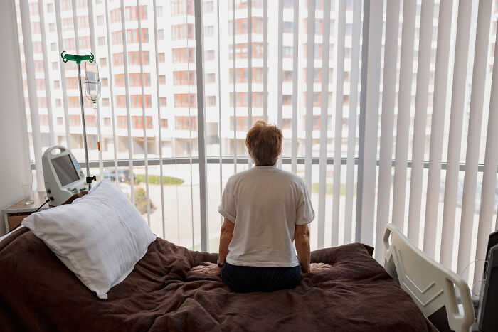 Elderly person sitting on a hospital bed near window blinds, evoking spooky tales of passing away and resurrection.