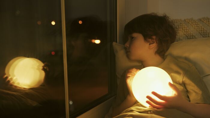 Child holding glowing orb while looking out window at night, reflecting on absurd things told growing up.
