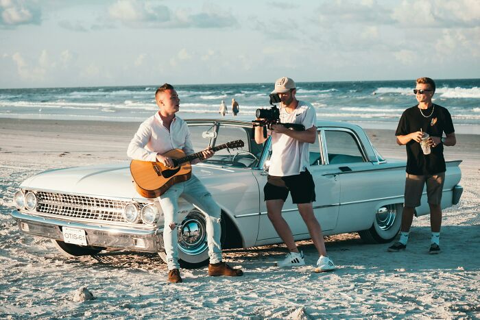 Musician playing guitar beside vintage car on the beach while a videographer films an iconic music video scene. Musician playing guitar beside vintage car on the beach while a videographer films an iconic music video scene.