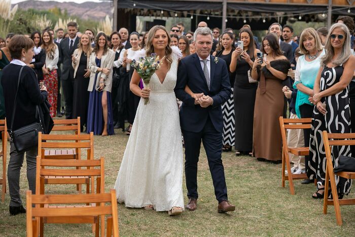 Bride walking down the aisle with a male escort at an outdoor wedding, surrounded by guests capturing the moment.