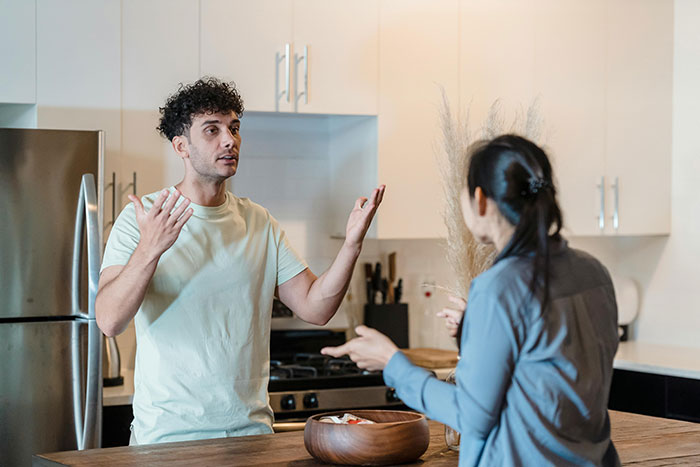 Man guarding box of olives at home, gesturing in frustration while woman cleans nearby in kitchen setting. Man guarding box of olives at home, gesturing in frustration while woman cleans nearby in kitchen setting.