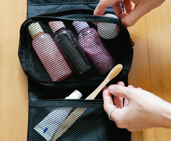 Hands organizing tiny travel bottles and a bamboo toothbrush inside a black mesh toiletry bag for a trip better.