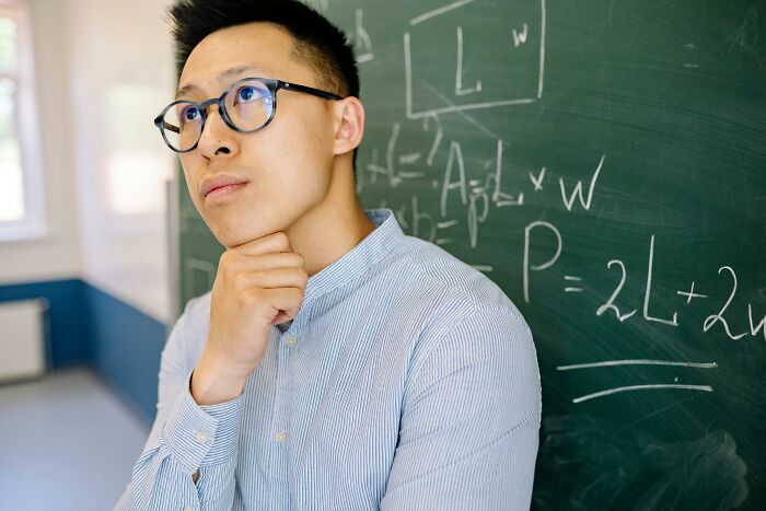 Young man wearing glasses thinking deeply in front of a chalkboard filled with math patterns and equations in a classroom setting Young man wearing glasses thinking deeply in front of a chalkboard filled with math patterns and equations in a classroom setting
