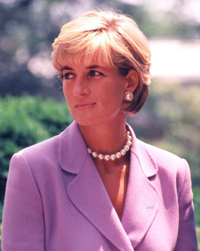 Princess Diana wearing a lavender suit and pearl necklace outdoors, looking thoughtfully to the side with a blurred background.