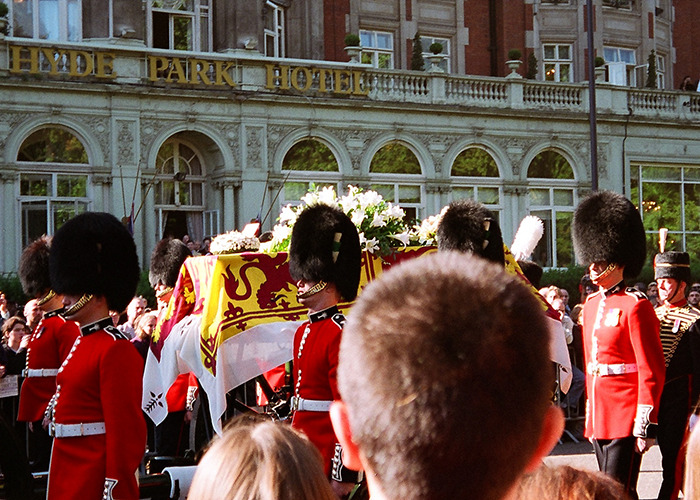 British guards in red uniforms carrying a royal coffin draped with a flag outside the Hyde Park Hotel during a royal procession.