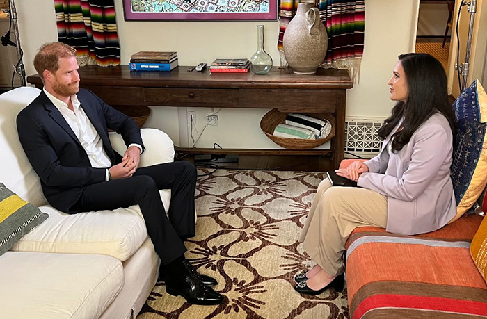 Prince Harry sitting on a white couch in a formal suit, talking to a woman in a beige blazer during an interview.