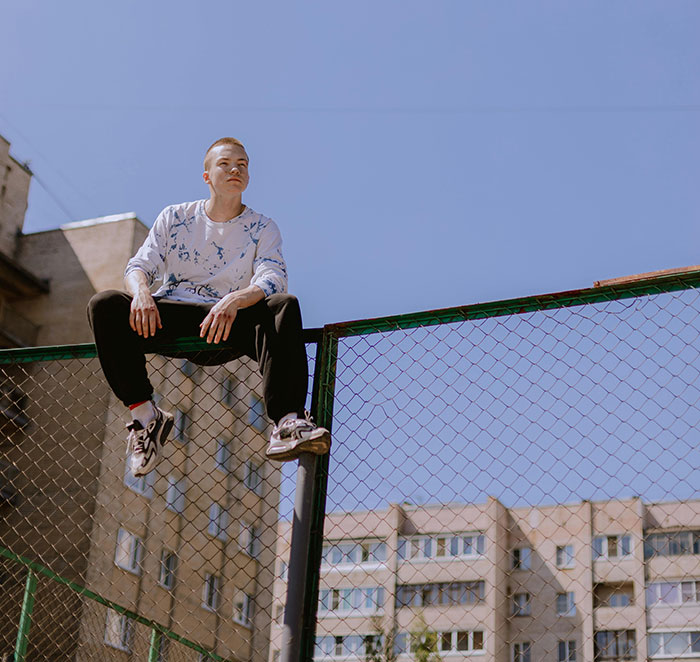 Young man taking huge risks sitting on top of a chain-link fence in an urban area under clear blue sky