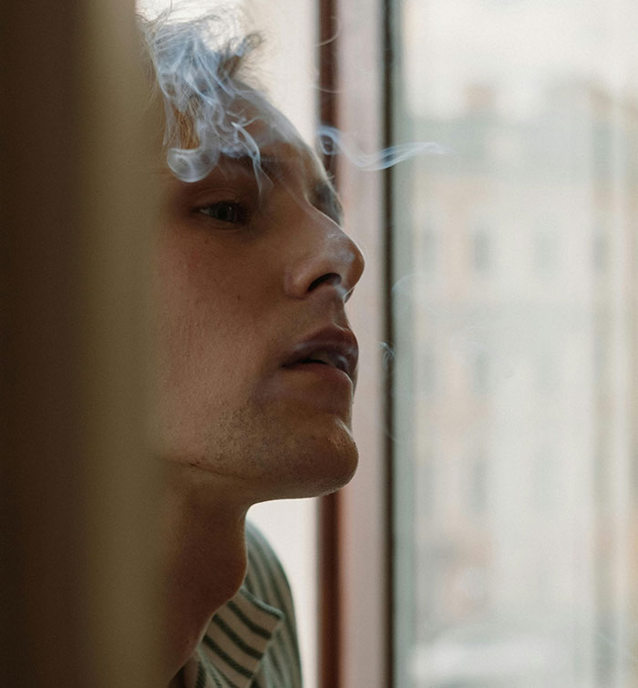 Young man risking health by smoking indoors near a window with smoke swirling around his face and head