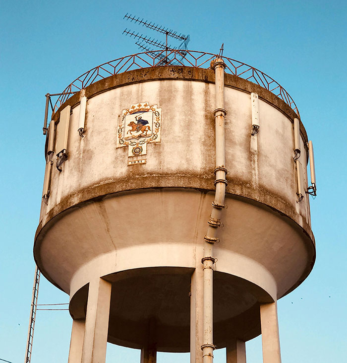 Old water tower with rusted pipes and antenna on top against clear blue sky, showing huge risks in unsafe conditions.