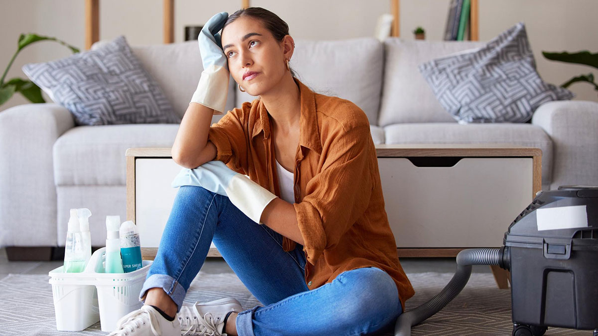 Woman wearing cleaning gloves sitting on floor looking frustrated boycotting chores at sexist parents' house with cleaning supplies nearby