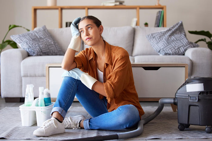 Young woman boycotts doing chores at sexist parents' house, waiting for her brothers to help clean the living room.