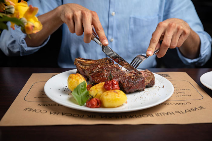 Person cutting steak with fork and knife, highlighting friend laughs off vegan’s warning about paying for his food. Person cutting steak with fork and knife, highlighting friend laughs off vegan’s warning about paying for his food.
