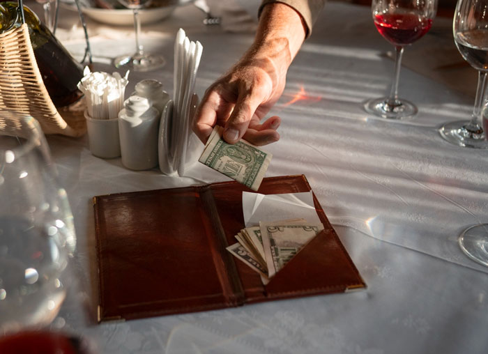 Man placing cash tip in check presenter at a restaurant table with wine glasses, reflecting friend’s GF ordering expensive meals. Man placing cash tip in check presenter at a restaurant table with wine glasses, reflecting friend’s GF ordering expensive meals.