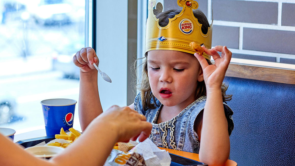 Young child wearing Burger King crown, eating fries at a restaurant, related to brotheru2019s picky kids in family trip story.