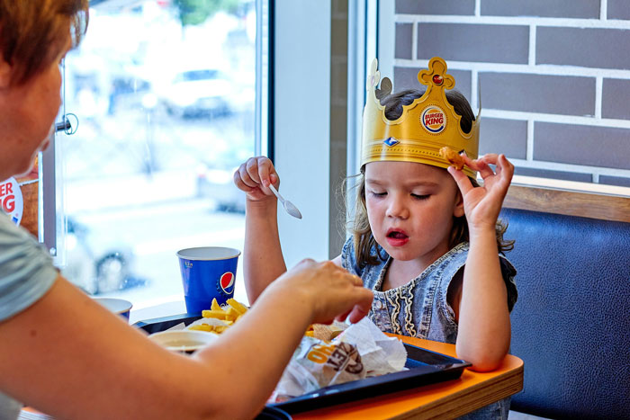 Child wearing Burger King crown eating fries at restaurant as adult reaches for food during a family meal outing.