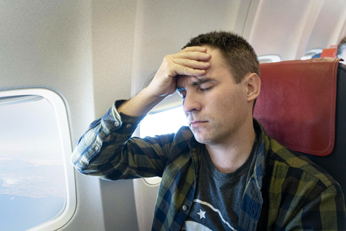 Man on plane with hand on forehead, appearing stressed while reflecting on life-changing trip after cancer battle. Man on plane with hand on forehead, appearing stressed while reflecting on life-changing trip after cancer battle.
