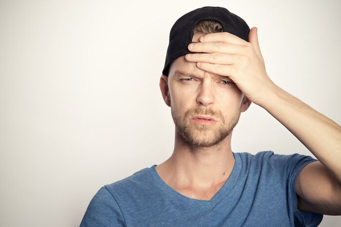 Young man in a blue shirt and black cap looks puzzled, holding his forehead contemplating mind-boggling beliefs.
