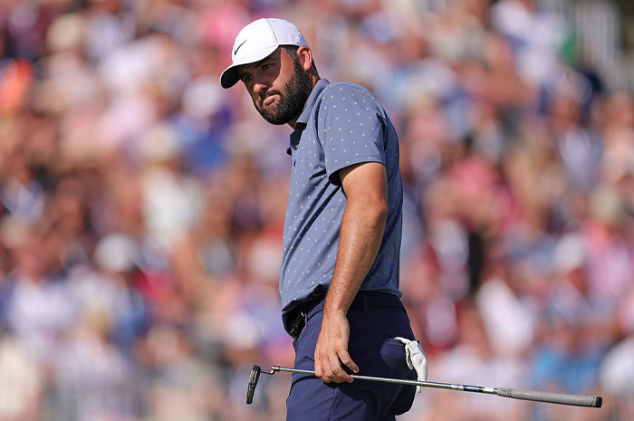 Scottie Scheffler focused on the green, holding a putter during a golf tournament with a crowded audience in the background.