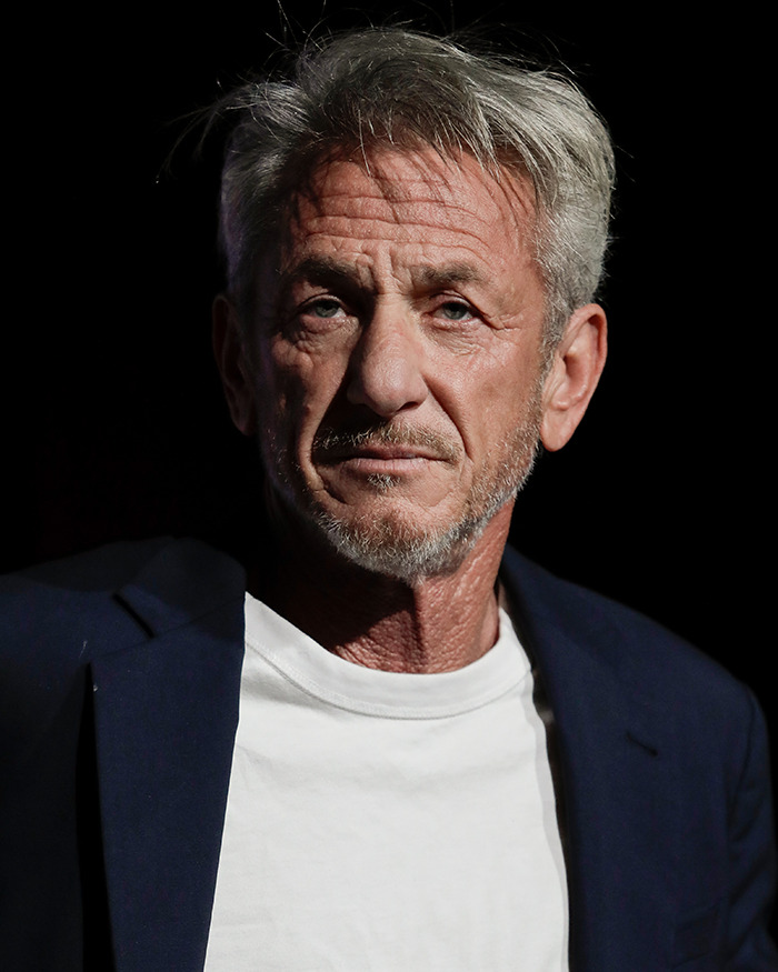 Sean Penn with a rough appearance, wearing a white shirt and dark blazer, photographed against a black background. Sean Penn with a rough appearance, wearing a white shirt and dark blazer, photographed against a black background.
