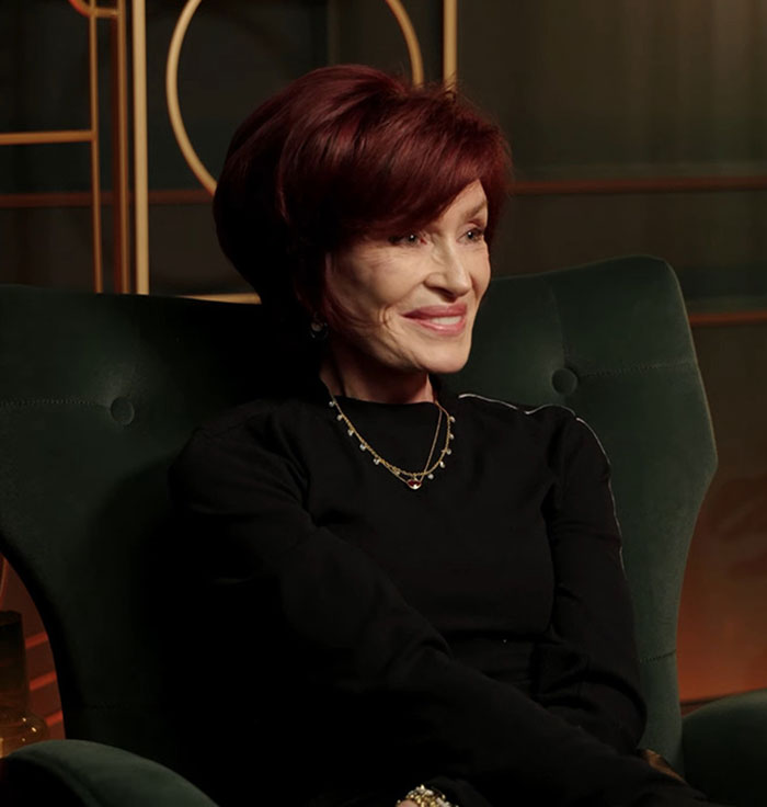 Sharon Osbourne sitting in a green chair wearing a black top and gold necklace, smiling during an interview indoors. Sharon Osbourne sitting in a green chair wearing a black top and gold necklace, smiling during an interview indoors.