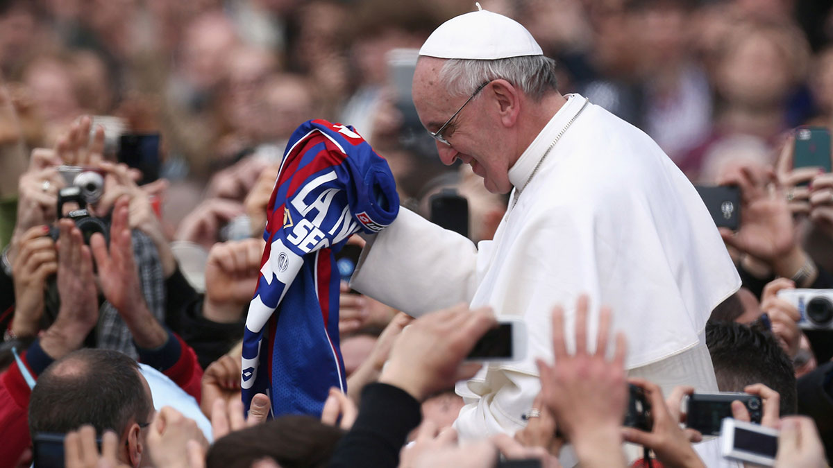 Pope Francis in white robes smiling and holding a sports jersey while surrounded by a crowd taking photos.