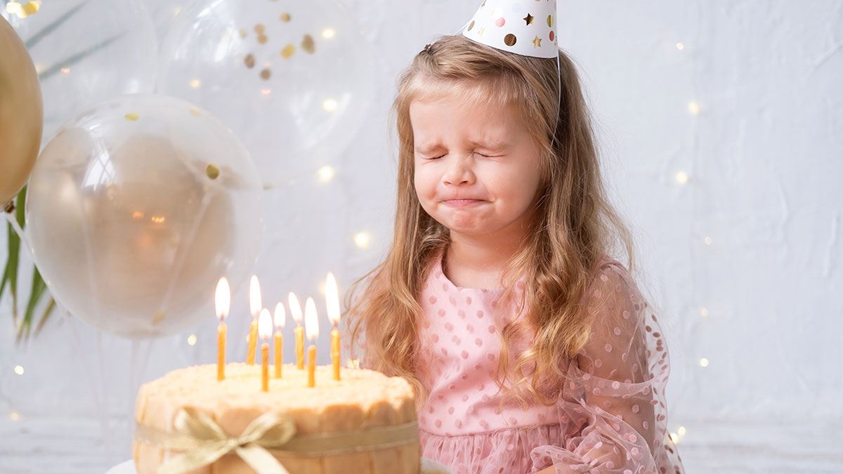 Young girl at her 11th birthday party looking upset with candles lit on the cake and party balloons in background