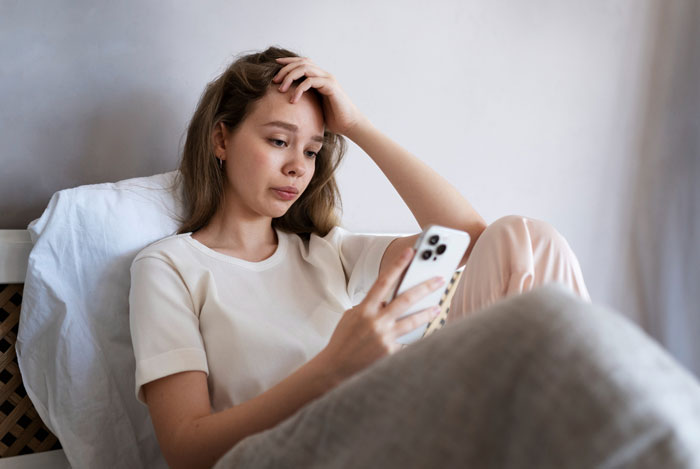 Sad 11-year-old girl sitting on bed, looking stressed while using smartphone during her birthday celebration.