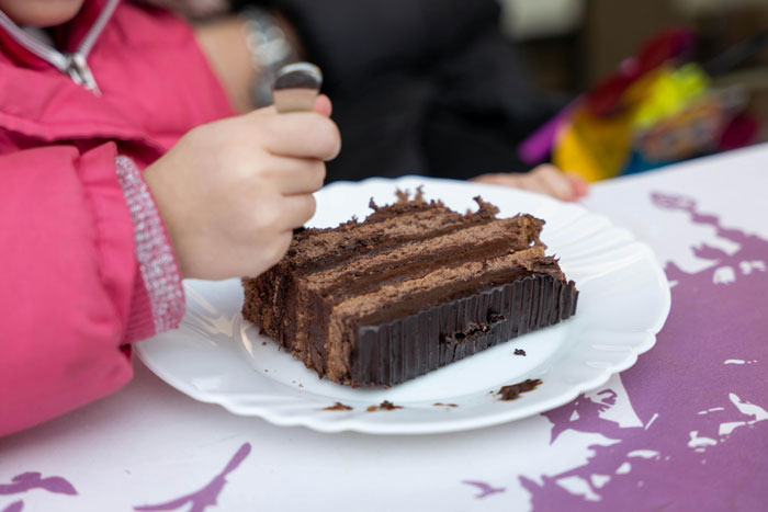Child in pink jacket holding fork, about to eat a piece of chocolate cake at a birthday gathering with visiting relatives.