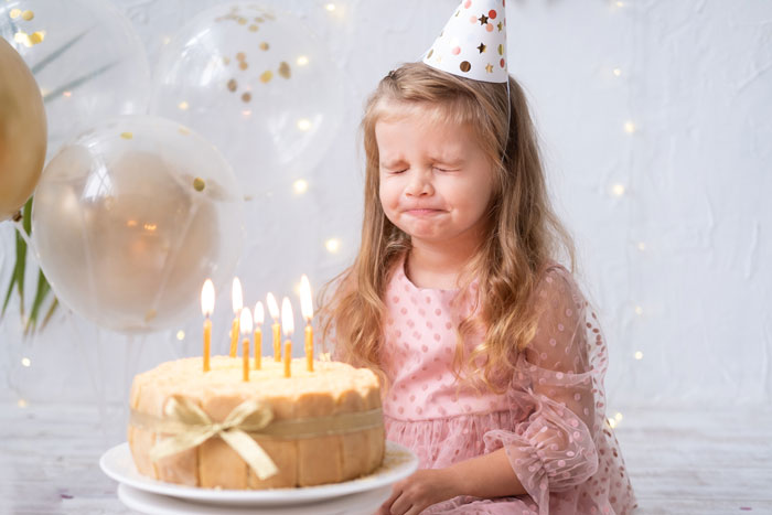 Young girl at her 11th birthday party looking upset near birthday cake with candles and balloons in background.