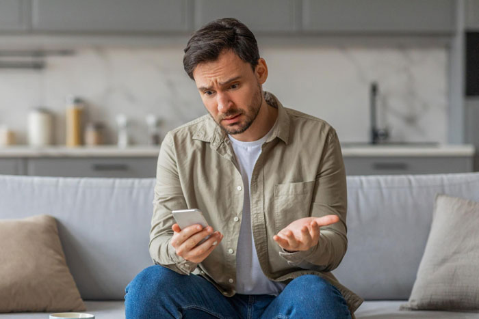 Man looking frustrated sitting on couch holding phone, representing girl’s 11th birthday visiting relatives demands.
