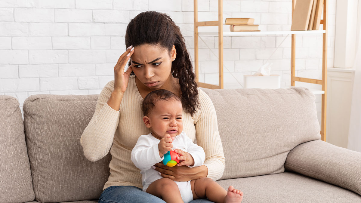 Woman babysitting crying baby niece and nephew, looking stressed and frustrated on a beige couch at home.