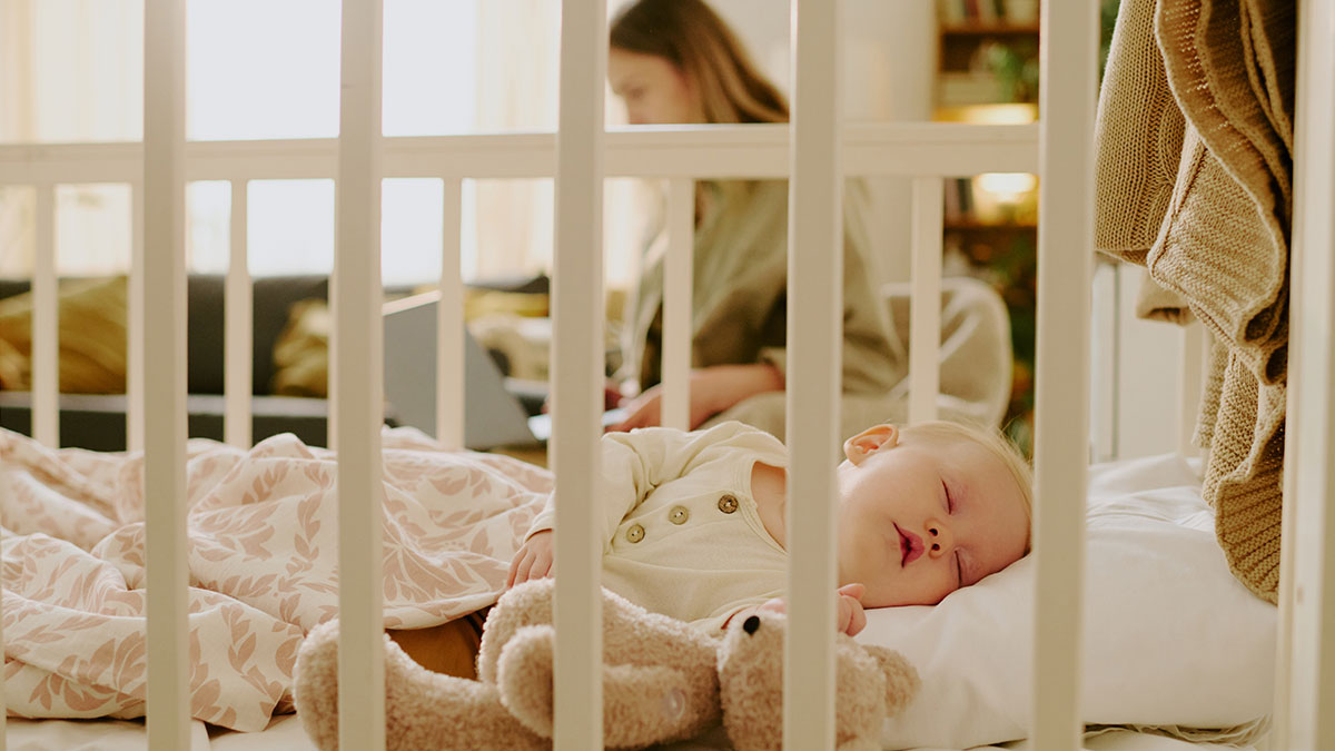 Baby falls asleep in crib with teddy bear nearby while family member works on laptop in background.