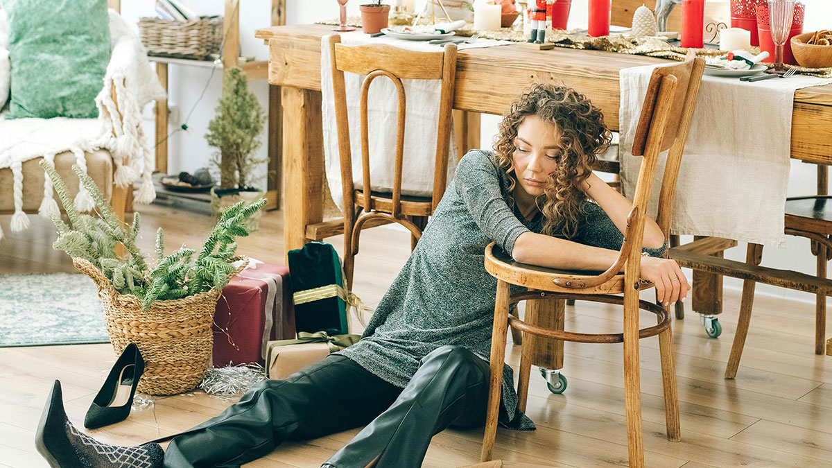 Woman appearing neglectful and tired sitting on the floor beside a dining table in a cluttered home setting