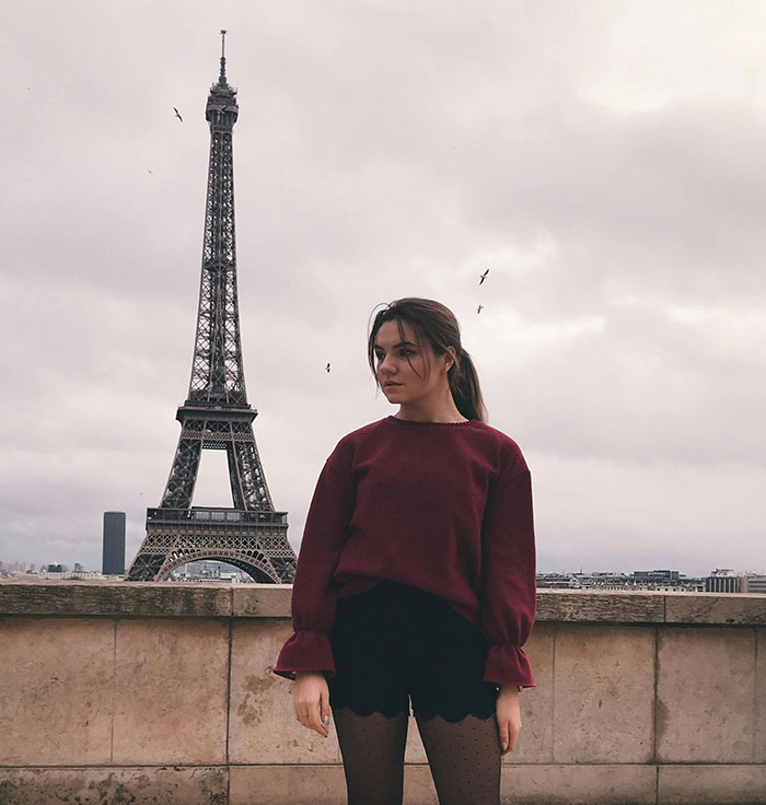 Young woman standing near Eiffel Tower in Paris, illustrating national stereotypes in a humorous travel context.