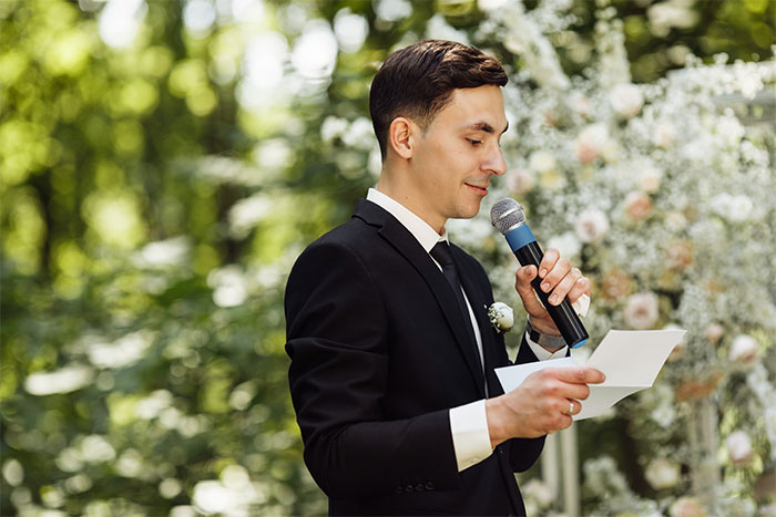 Man in a black suit holding a microphone and paper, speaking at a best friend's wedding during a proposal moment. Man in a black suit holding a microphone and paper, speaking at a best friend's wedding during a proposal moment.