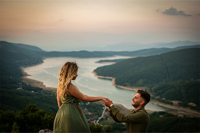 Couple overlooking a scenic river as the man kneels to propose, capturing a moment of a boyfriend’s proposal at a wedding. Couple overlooking a scenic river as the man kneels to propose, capturing a moment of a boyfriend’s proposal at a wedding.